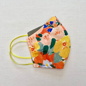 May include: A reusable face mask with a floral print featuring orange, yellow, pink, blue, and green flowers on a white background. The mask has yellow elastic straps.