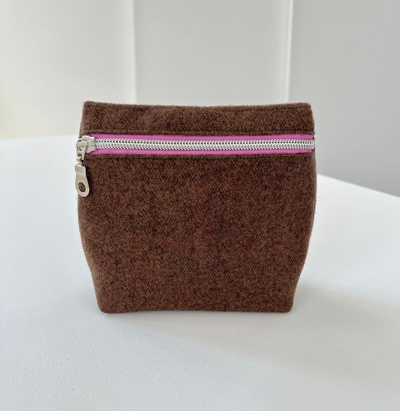 May include: A brown felt zippered pouch with a pink zipper pull. The pouch is sitting on a white surface.