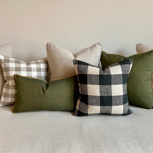 May include: A set of five throw pillows with a variety of patterns. The pillows are arranged on a light grey sofa. The pillows include a black and white buffalo check pattern, a green solid pillow with a yellow trim, and two solid light grey pillows.
