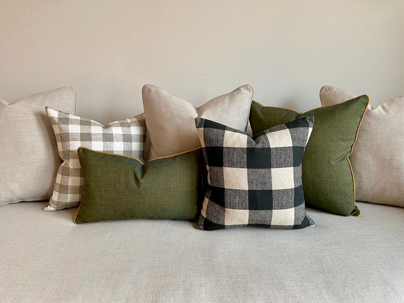 May include: A set of five throw pillows with a variety of patterns. The pillows are arranged on a light grey sofa. The pillows include a black and white buffalo check pattern, a green solid pillow with a yellow trim, and two solid light grey pillows.