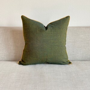May include: A square, olive green decorative pillow with a gold-colored trim. The pillow is made of a textured fabric and is placed on a light gray sofa. The pillow has a simple, elegant design.