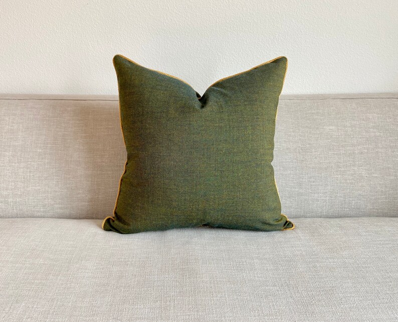 May include: A square, olive green decorative pillow with a gold-colored trim. The pillow is made of a textured fabric and is placed on a light gray sofa. The pillow has a simple, elegant design.