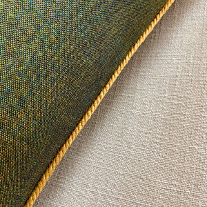 May include: Close-up of a green fabric cushion with a gold trim. The cushion is lying on a beige fabric surface.