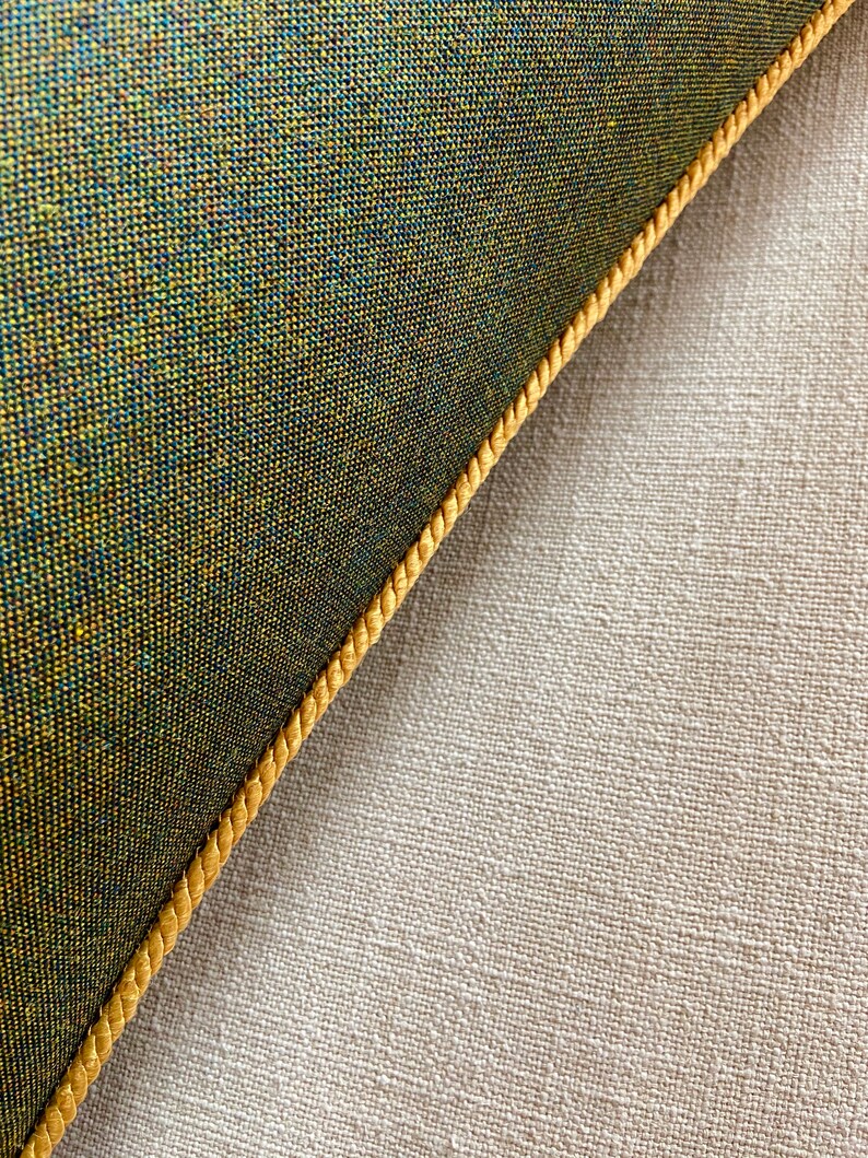 May include: Close-up of a green fabric cushion with a gold trim. The cushion is lying on a beige fabric surface.