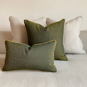 May include: Four decorative throw pillows in a neutral color palette. Two pillows are a light beige, and two are a deep olive green with gold piping. The pillows are arranged on a light gray sofa.