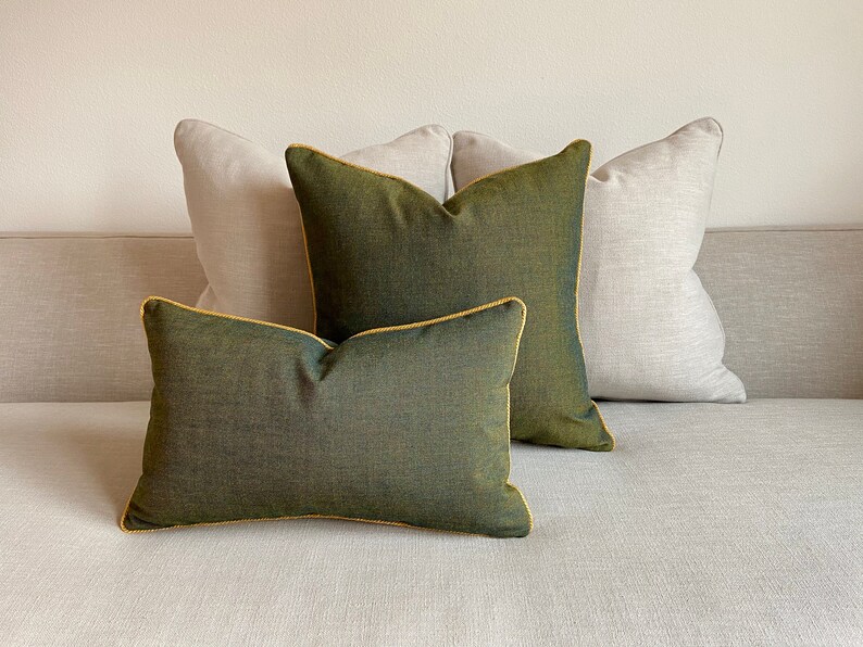 May include: Four decorative throw pillows in a neutral color palette. Two pillows are a light beige, and two are a deep olive green with gold piping. The pillows are arranged on a light gray sofa.