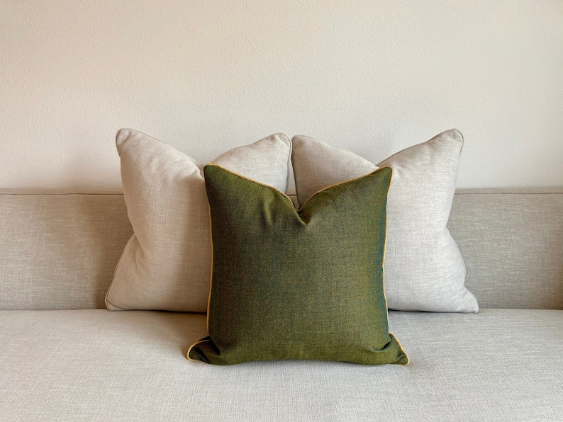 May include: Three decorative throw pillows on a light-colored sofa. The center pillow is a deep green with gold trim, flanked by two neutral-toned pillows. The pillows add a touch of color and texture to the neutral sofa.