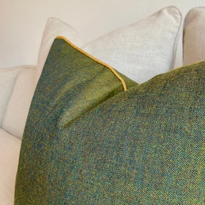 May include: Close-up of a decorative pillow with a textured green and blue fabric. The pillow has a gold-colored trim and is placed on a white sofa. The pillow's fabric has a woven appearance.