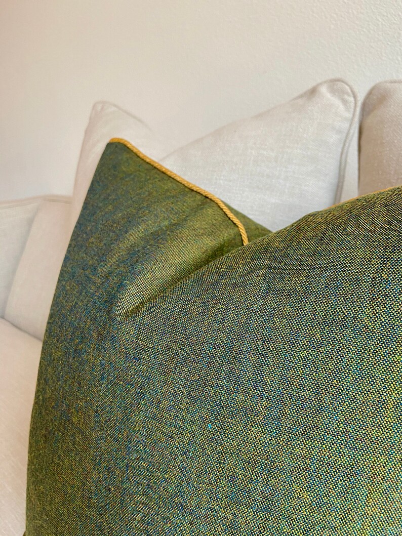 May include: Close-up of a decorative pillow with a textured green and blue fabric. The pillow has a gold-colored trim and is placed on a white sofa. The pillow's fabric has a woven appearance.
