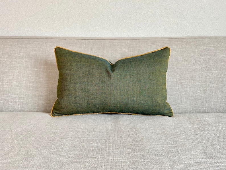 May include: A dark green cushion with a gold trim. The cushion is sitting on a light grey sofa.