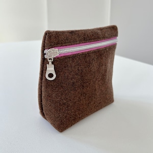 May include: A brown felt zippered pouch with a pink zipper pull. The pouch is lying on a white surface.