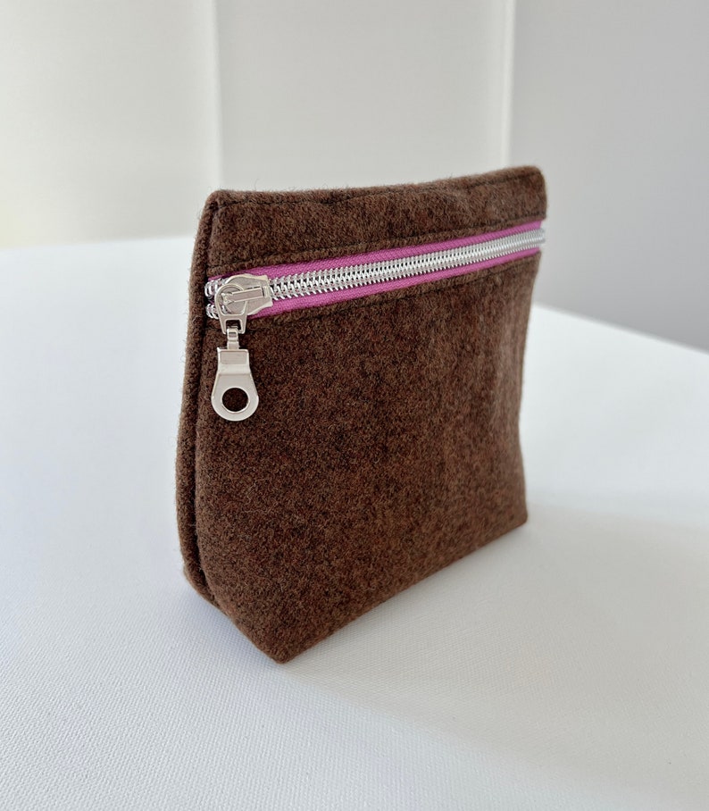 May include: A brown felt zippered pouch with a pink zipper pull. The pouch is lying on a white surface.