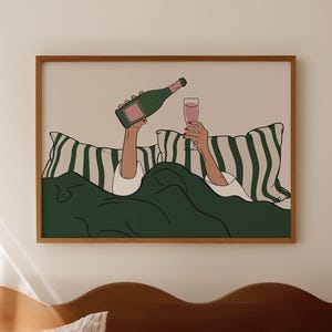Pode incluir: Uma impressão artística emoldurada retrata uma pessoa na cama segurando uma garrafa de champanhe e um copo de líquido rosa. A obra de arte apresenta uma garrafa verde, um copo rosa e um cobertor verde. O fundo inclui travesseiros listrados e uma parede neutra.