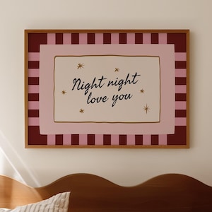 May include: A framed print with the text "Night night love you" in a bedroom setting. The print has a pink and burgundy color scheme with a gold frame. The bed has striped bedding and a wooden headboard.