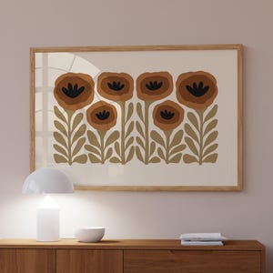 May include: A mid-century modern style sideboard made of wood with two doors and three drawers. The sideboard is light brown and has a white lamp and a framed print of brown flowers on a white background above it.