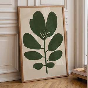 May include: A framed botanical print with a stylized green flower and leaves on a cream background. The artwork is displayed in a natural wood frame. The print leans against a white wall and wooden floor.