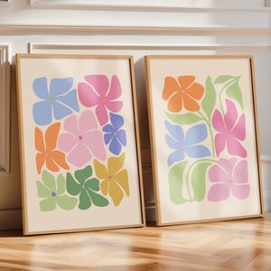 K&ouml;nnte beinhalten: Zwei gerahmte Kunstdrucke mit farbenfrohen, abstrakten Blumendesigns. Die Blumen sind in Blau-, Rosa-, Orange-, Gr&uuml;n- und Gelbt&ouml;nen auf cremefarbenem Hintergrund gehalten. Die Rahmen sind helles Holz, und das Kunstwerk lehnt an einer wei&szlig;en Wand.