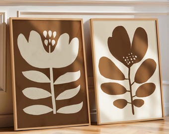 Op de afbeelding: Twee ingelijste kunstprints met bloemen. Eén print toont een grote witte bloem op een bruine achtergrond. De andere heeft een bruine bloem op een crèmekleurige achtergrond. Beide zitten in houten frames en leunen tegen een witte muur.
