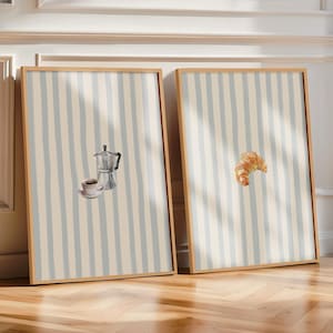 Könnte beinhalten: Zwei gerahmte Kunstdrucke mit Kaffeemotiv. Ein Druck zeigt eine Kaffeemaschine und eine Tasse Kaffee, der andere ein Croissant. Beide haben einen hellblauen und beigefarbenen gestreiften Hintergrund und einen Holzrahmen.