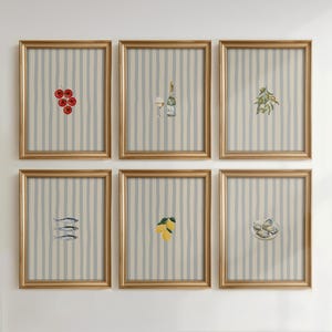 May include: A set of six framed wall art prints, each featuring a centered illustration of food or drink items including tomatoes, wine, olives, fish, lemons, and oysters, set against a light blue and white vertical striped background in gold frames.