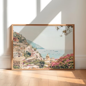 May include: A framed photograph of a coastal town with colorful buildings on a hillside, overlooking a blue ocean with boats. The photo is in a light wood frame and has a white background.
