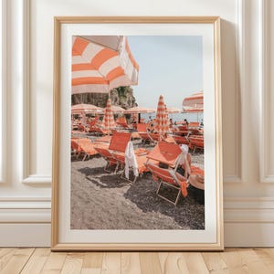 May include: A framed print of a beach scene with orange and white striped umbrellas and lounge chairs. The beach is filled with people enjoying the sun. The print is in a wooden frame and is displayed against a white wall.