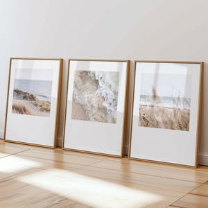 May include: Three framed art prints with coastal themes. Each print is set in a light brown wooden frame with a white border. The images show a beach with waves, sand, and grasses. The frames are leaning against a white wall on a wooden floor.
