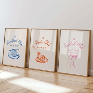 May include: Three framed prints with a light brown background and a wooden frame. The prints are titled "The Breakfast Club", "The Pasta Club", and "The Cocktail Club". Each print features a stylized illustration of food or a drink. The Breakfast Club print features a blue illustration of a stack of pancakes with syrup. The Pasta Club print features an orange illustration of a plate of spaghetti with a fork. The Cocktail Club print features a pink illustration of a martini glass.