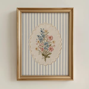 K&ouml;nnte beinhalten: Ein gerahmtes Blumenstickerei. Das Kunstwerk zeigt einen Strau&szlig; aus rosa, blauen und gelben Blumen auf einem ovalen, cremefarbenen Hintergrund mit einem gewellten Rand. Der Hintergrund ist ein hellblau-wei&szlig; gestreifter Stoff, der in einem goldfarbenen Rahmen eingefasst ist.