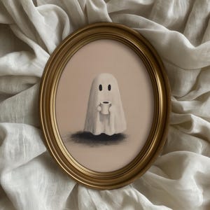 May include: An oval, gold-framed artwork featuring a whimsical ghost holding a small cup. The ghost is white with two black eyes, set against a pale pink background. The artwork has a soft, illustrative style, with a dark shadow beneath the ghost.
