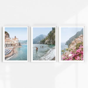 May include: Three framed prints of coastal scenes. The first print shows a white building with a blue roof on a rocky cliff overlooking a turquoise ocean. The second print shows a rocky cliff with a turquoise ocean and people swimming. The third print shows a rocky cliff with a turquoise ocean and pink flowers in the foreground.
