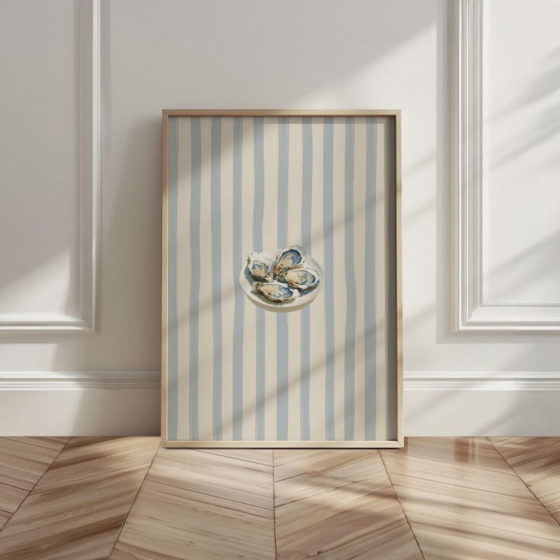 May include: A framed print featuring a watercolor illustration of oysters on a white plate. The background has vertical stripes in light blue and cream. The frame is a light wood color. The artwork is displayed against a white wall and wooden floor.