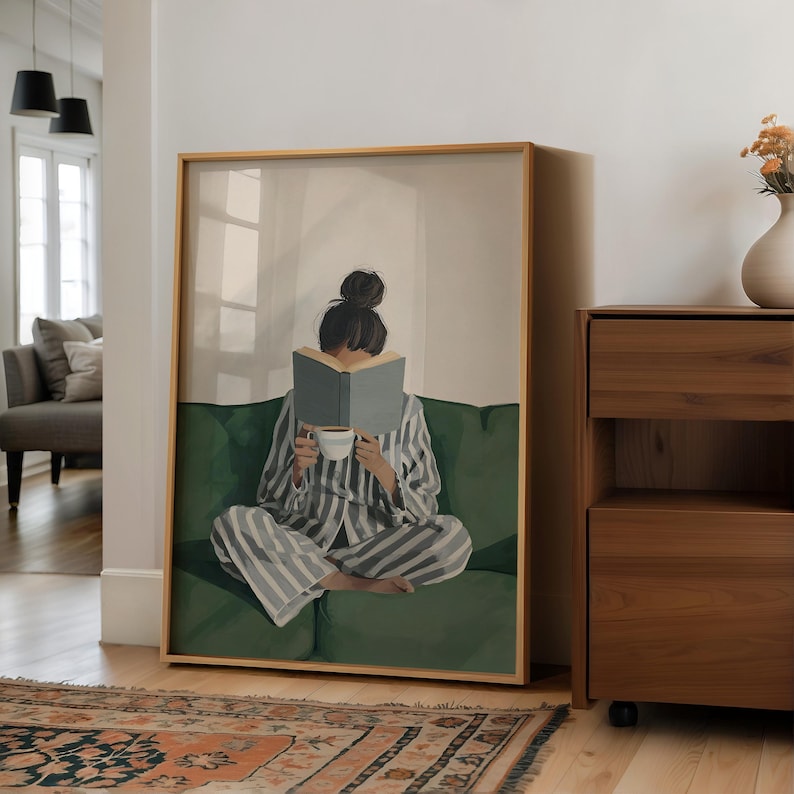 May include: Framed art print featuring a person in striped pajamas reading a book while seated on a green sofa. The person holds a cup of coffee. The artwork has a light wood frame and is displayed in a well-lit room with a rug.