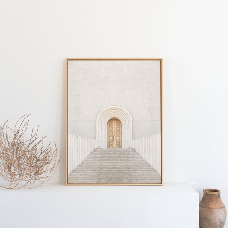 May include: A framed print of a minimalist architectural photograph. The image features a white building with a set of double doors and a staircase leading up to them. The doors are a light wood color and the staircase is made of white stone.
