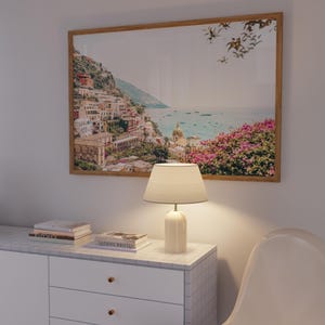 May include: A framed print of a coastal Italian town hangs above a white cabinet with a lamp and books. The print features colorful buildings on a hillside overlooking the sea. A white lamp with a beige shade sits on the cabinet.