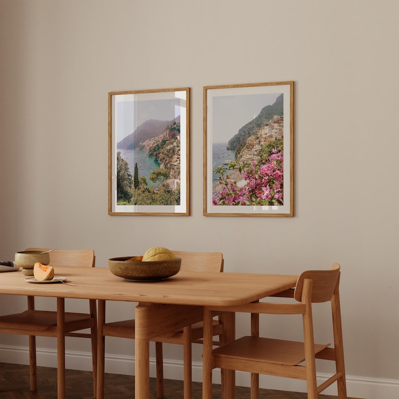 May include: Two framed landscape photographs of a coastal town with a view of the ocean and mountains. The photographs are hanging on a beige wall above a wooden dining table with four chairs.