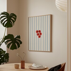May include: A framed art print showing a bunch of red cherries set against a background of vertical blue and cream stripes. The artwork is presented in a light-coloured wooden frame, adding a touch of natural warmth to the design.