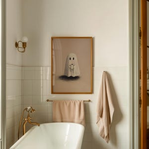 May include: A framed art print featuring a cartoon ghost holding a cup, set against a neutral background. The artwork is in a gold frame and hangs above a towel rack with a peach-colored towel in a bathroom setting.