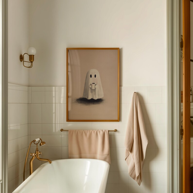 May include: A framed art print featuring a cartoon ghost holding a cup, set against a neutral background. The artwork is in a gold frame and hangs above a towel rack with a peach-colored towel in a bathroom setting.