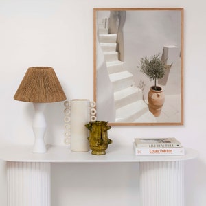 K&ouml;nnte beinhalten: Wei&szlig;er Konsolentisch mit einer wei&szlig;en Lampe mit einem braunen geflochtenen Schirm, einer wei&szlig;en Keramikvase mit Ringdesign und einer gr&uuml;nen Keramikvase. &Uuml;ber dem Tisch h&auml;ngt ein gerahmtes Bild eines wei&szlig;en Treppenhauses mit einem Topf-Olivenbaum. Zwei B&uuml;cher liegen auf dem Tisch, eines mit dem Text "Live Beautiful" und das andere mit dem Text "Louis Vuitton."
