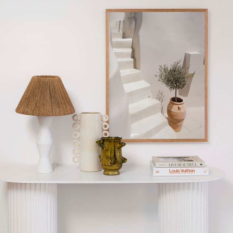 K&ouml;nnte beinhalten: Wei&szlig;er Konsolentisch mit einer wei&szlig;en Lampe mit einem braunen geflochtenen Schirm, einer wei&szlig;en Keramikvase mit Ringdesign und einer gr&uuml;nen Keramikvase. &Uuml;ber dem Tisch h&auml;ngt ein gerahmtes Bild eines wei&szlig;en Treppenhauses mit einem Topf-Olivenbaum. Zwei B&uuml;cher liegen auf dem Tisch, eines mit dem Text "Live Beautiful" und das andere mit dem Text "Louis Vuitton."