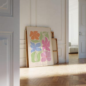 K&ouml;nnte beinhalten: Ein gerahmter Kunstdruck mit einem Blumendesign in Orange, Rosa, Blau und Gr&uuml;n. Das Kunstwerk wird vor anderen gerahmten St&uuml;cken in einem Raum mit Holzboden pr&auml;sentiert. Der Druck wird vor einem neutralen Hintergrund angezeigt.