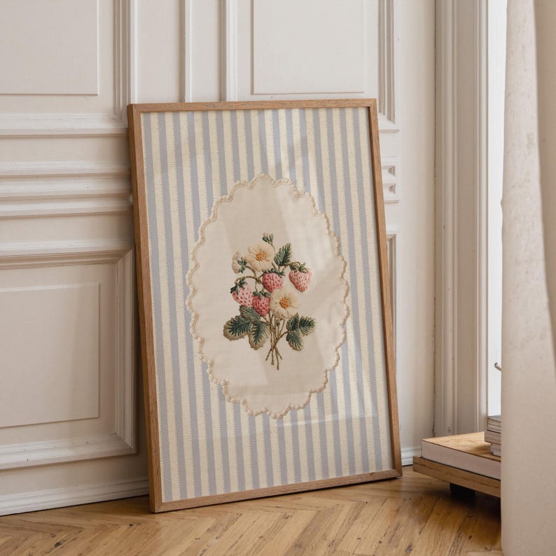 May include: A framed artwork featuring a vintage-style strawberry and flower bouquet. The artwork is set against a striped background of light blue and white. The frame is made of light-colored wood, and the artwork is leaning against a wall.
