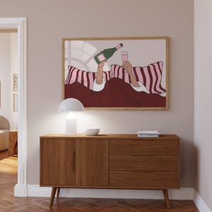 K&ouml;nnte beinhalten: Ein gerahmter Kunstdruck zeigt eine Person im Bett, die Champagner in ein Glas gie&szlig;t. Das Kunstwerk zeigt eine gr&uuml;ne Champagnerflasche, ein rosa Glas und rosa-wei&szlig; gestreifte Kissen. Das Farbschema ist rosa, rot und wei&szlig;.