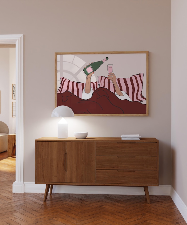 K&ouml;nnte beinhalten: Ein gerahmter Kunstdruck zeigt eine Person im Bett, die Champagner in ein Glas gie&szlig;t. Das Kunstwerk zeigt eine gr&uuml;ne Champagnerflasche, ein rosa Glas und rosa-wei&szlig; gestreifte Kissen. Das Farbschema ist rosa, rot und wei&szlig;.