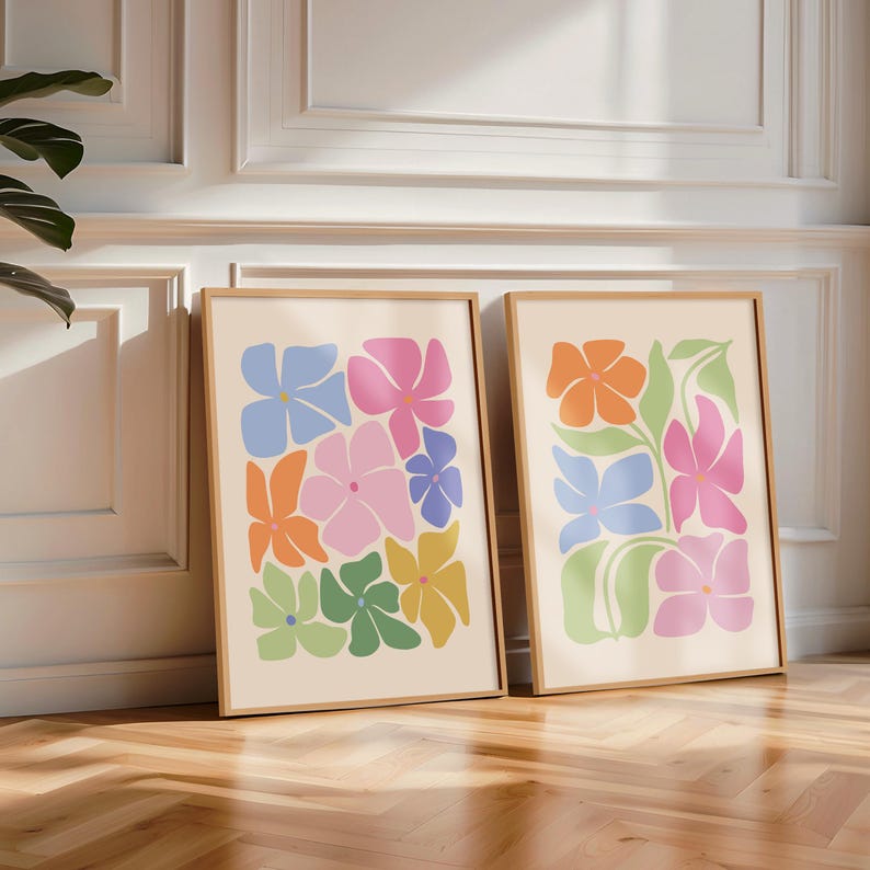 K&ouml;nnte beinhalten: Zwei gerahmte Kunstdrucke mit farbenfrohen, abstrakten Blumendesigns. Die Blumen sind in Blau-, Rosa-, Orange-, Gr&uuml;n- und Gelbt&ouml;nen auf cremefarbenem Hintergrund gehalten. Die Rahmen sind helles Holz, und das Kunstwerk lehnt an einer wei&szlig;en Wand.