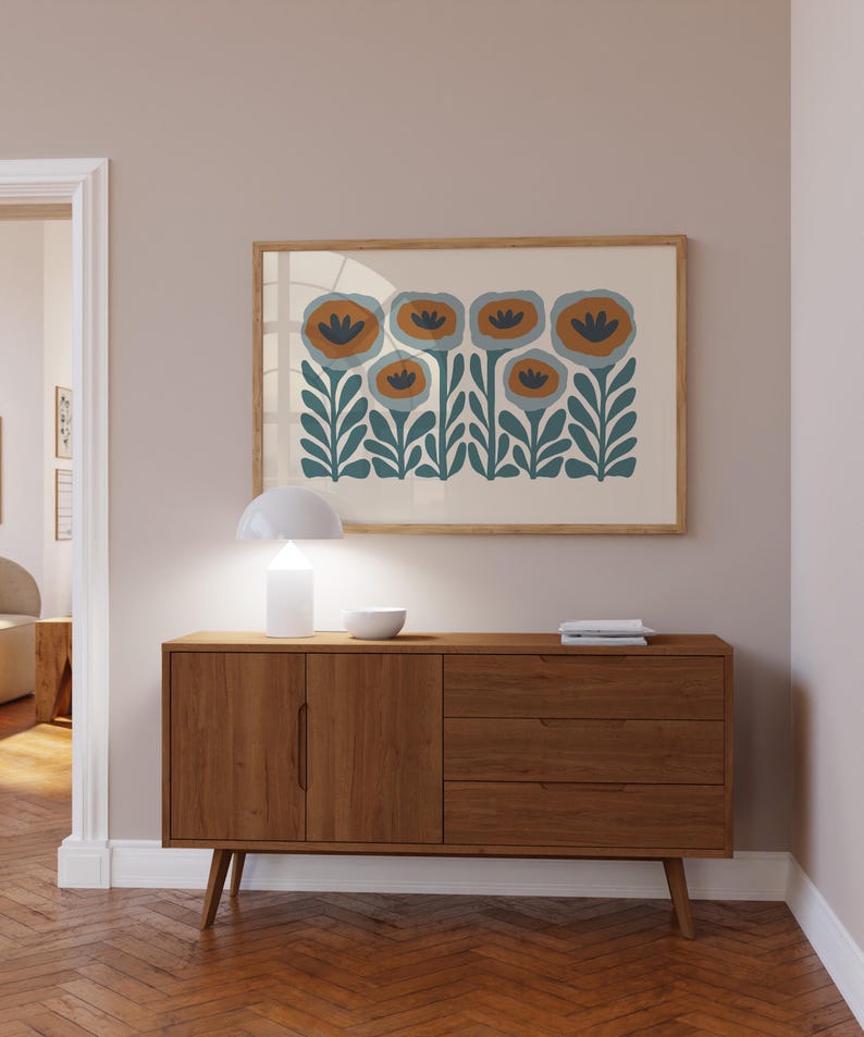May include: A framed floral art print with orange and blue flowers hangs above a wooden mid-century modern style sideboard. A white lamp and bowl sit on top of the sideboard. The room has a neutral color palette with a herringbone wood floor.