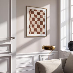 May include: A framed abstract print with a checkered pattern in shades of brown and white. The print is hanging on a white wall in a room with a couch and a lamp.