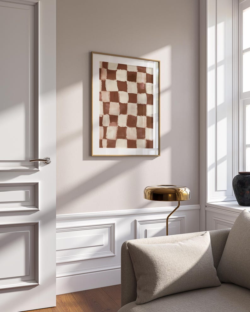 May include: A framed abstract print with a checkered pattern in shades of brown and white. The print is hanging on a white wall in a room with a couch and a lamp.