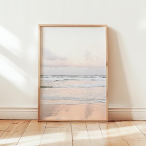 Op de afbeelding: Een ingelijste foto van een strandscène met golven die breken op de kust. De lucht is zacht roze en paars, en het zand is licht beige.